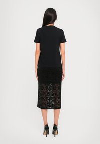Woman with long dark hair wearing a black short-sleeve top, black floral lace midi skirt, and black high-heeled shoes, standing facing away.