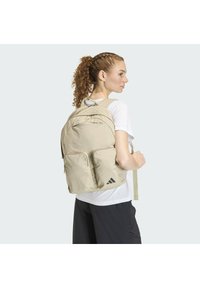 Mochila de tela beige con patrón de cuadrícula, múltiples compartimentos, correas acolchadas para los hombros y detalles de logo en negro.