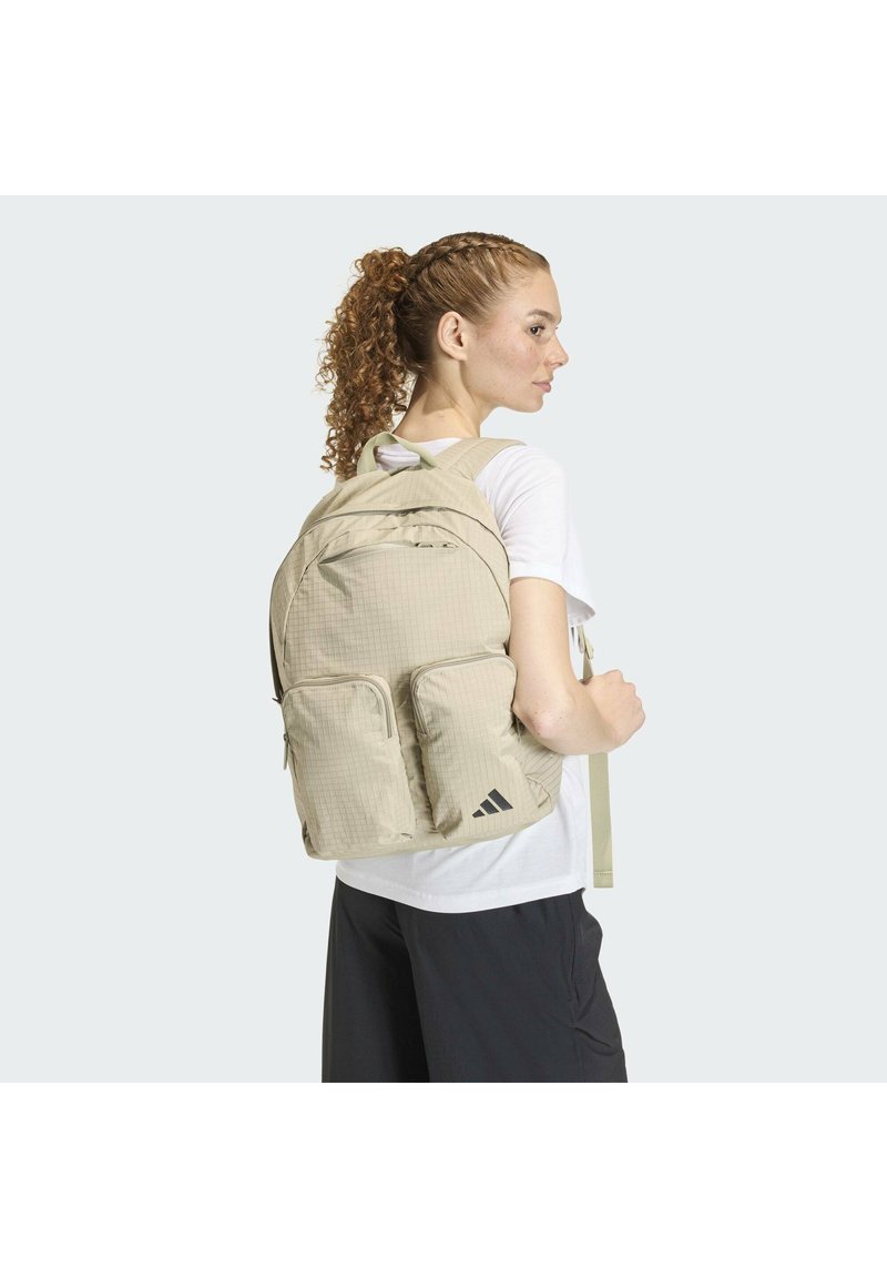 Mochila de tela beige con patrón de cuadrícula, múltiples compartimentos, correas acolchadas para los hombros y detalles de logo en negro.