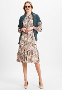 Bloemrijke jurk met een gerimpelde zoom in roze, beige en groene tinten. Gehaakte teal omslagdoek over de schouders. Ronde zonnebril en nude hoge hakken.