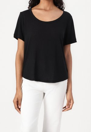 Femme portant un t-shirt noir à manches courtes et col rond et un pantalon blanc, montrée des épaules aux hanches sur un fond uni.