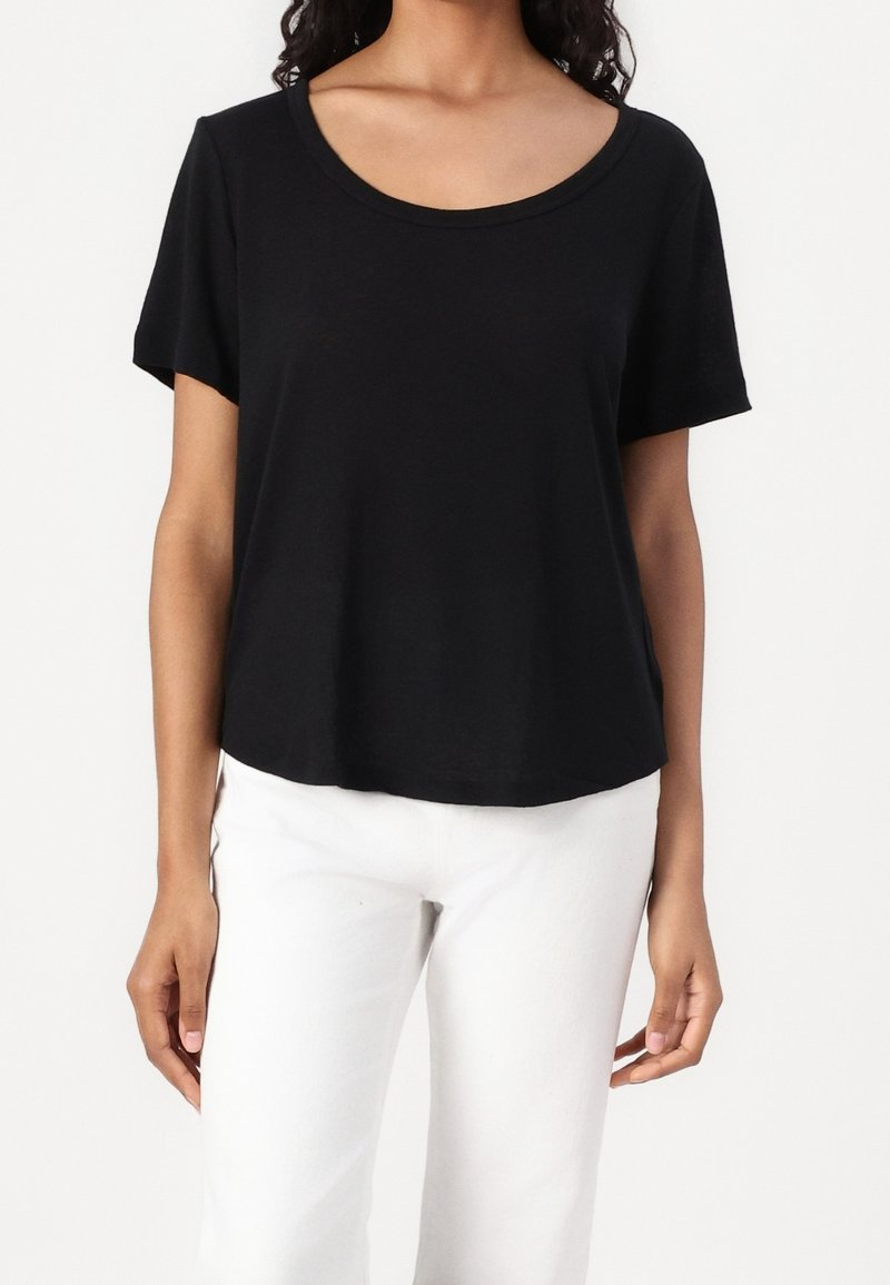 Femme portant un t-shirt noir à manches courtes et col rond et un pantalon blanc, montrée des épaules aux hanches sur un fond uni.