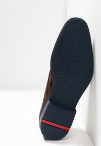 Chaussure en daim marron avec une semelle en caoutchouc noir texturé présentant un motif hexagonal et un accent rouge à l'arrière.
