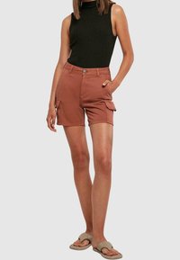 Shorts cargo de couleur rouille, en coton, avec poches latérales et une fermeture à boutons sur l'avant. Associés à un haut noir sans manches.