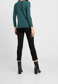 Turkusowa bluzka z długim rękawem o luźnym kroju, zestawiona z wąskimi czarnymi spodniami i czarnymi botkami na obcasie, widok od tyłu.