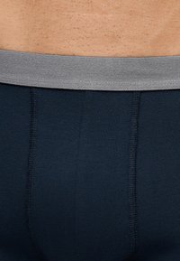 Nærbillede af en persons nedre torso iført mørkeblå undertøj med en grå talje, der viser hud- og stoffteksturer.