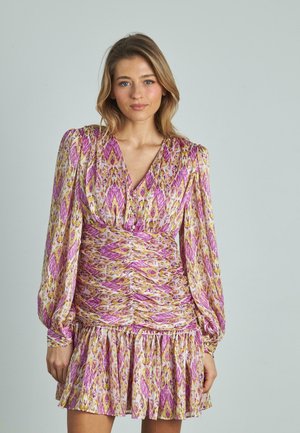 Mujer de pie contra un fondo liso, vistiendo un vestido de manga larga, con fruncidos, de colores rosa, amarillo y blanco, con escote en V y dobladillo con volantes.