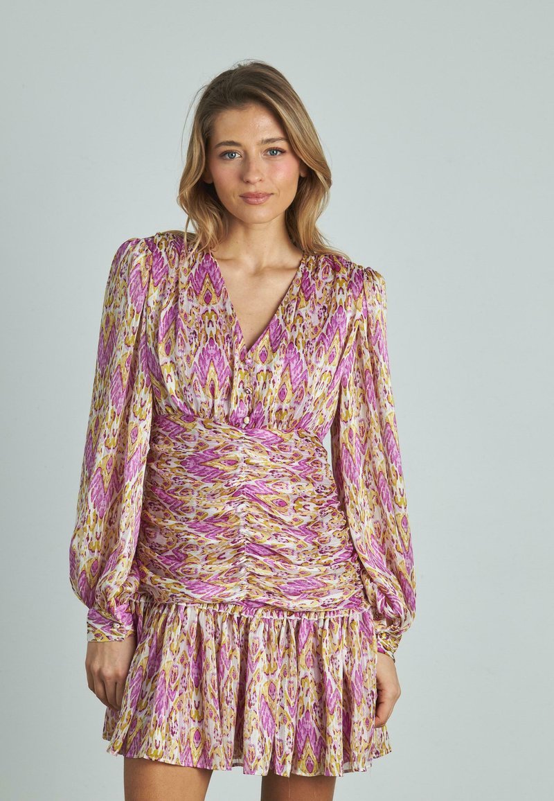 Mujer de pie contra un fondo liso, vistiendo un vestido de manga larga, con fruncidos, de colores rosa, amarillo y blanco, con escote en V y dobladillo con volantes.