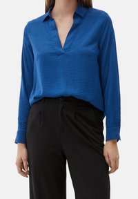 Blusa blu a maniche lunghe con colletto, scollatura a V e texture stropicciata, abbinata a pantaloni neri a vita alta. Accenti a bottoni visibili sui polsini.