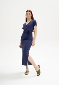 Marineblaue Wickelkleid mit kurzen Ärmeln, tailliert und mit seitlichem Schlitz. Aus weichem Stoff gefertigt, kombiniert mit olivgrünen Blockabsatzsandalen.