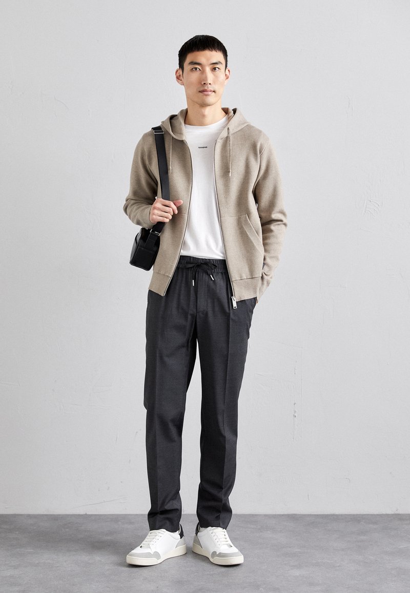 Hellbrauner Zip-Hoodie über einem weißen T-Shirt, kombiniert mit dunkelgrauen Hosen und weißen Sneakers. Schwarze Tasche über der Schulter.