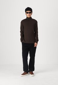 Pulover turtleneck de culoare maro închis, cu dungi, asortat cu pantaloni din catifea neagră și adidași maro, completat cu ochelari de soare negri.