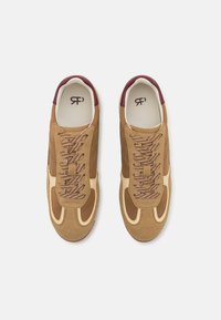Braune Wildleder-Sneaker mit cremefarbenen Akzenten, ausgestattet mit einem bordeauxroten Fersenrand und strukturierten Schnürsenkeln. Die Fußbett hat ein schwarzes Logo aufgedruckt.