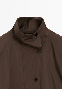 Manteau marron à col montant avec trois boutons noirs et coutures visibles aux épaules et sur le panneau avant sur fond blanc.