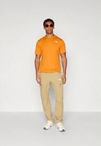 Oransje t-skjorte med korte ermer og beige cargobukser, begge med logo. Modellen har på seg solbriller og lyse sportssko.