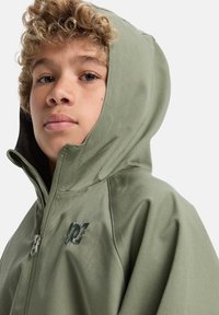 Oliivinvihreä vetoketjullinen huppari, jossa on kestävä kangas, näkyvä vetoketju ja musta logo yksityiskohtana rinnassa. Huppu on muotoiltu istuvaksi.