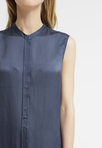 Marineblaue ärmellose Bluse aus strukturiertem Stoff, mit Knopfleiste vorne und klassischem Kragen, der einen dezenten Glanz aufweist.