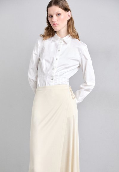 Camicia bianca a maniche lunghe con bottoni e colletto, abbinata a una gonna avvolgente crema fluida con legatura laterale. Design semplice e moderno.