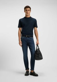 Navy short-sleeve t-shirt, fitted design; slim dark jeans; black leather loafers; holding a textured black handbag with shoulder strap.