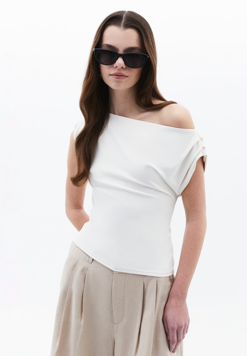 Top bianco off-the-shoulder con dettaglio in tessuto drappeggiato, maniche corte arrotolate, abbinato a pantaloni beige a vita alta con pieghe.