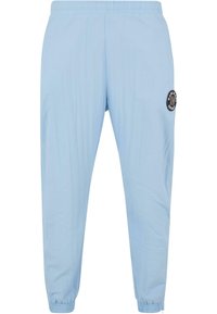 Light blue athletic pants with an elastic waistband, tapered fit, and a black circular logo patch on the left thigh. Smooth texture.