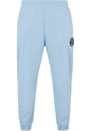 Light blue athletic pants with an elastic waistband, tapered fit, and a black circular logo patch on the left thigh. Smooth texture.