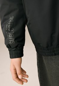 Zwarte bomberjack met reflecterende tekst "SUBSCRIBE," voorzien van elastische manchetten en een gladde, gestructureerde stof.