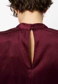 Bordeaux blouse met een sleutelgatopening aan de achterkant, subtiele gekreukte textuur en een mandaryn kraag met twee knoopsluitingen.