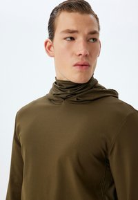 Camiseta deportiva de manga larga en verde oliva con cuello alto, textura suave y un diseño ajustado. Presenta un sutil detalle en el panel lateral.