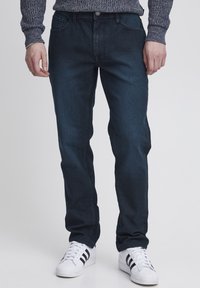 Jeans in denim blu scuro con una vestibilità dritta classica, cinque tasche e zip. Indossati con sneakers bianche con strisce nere.