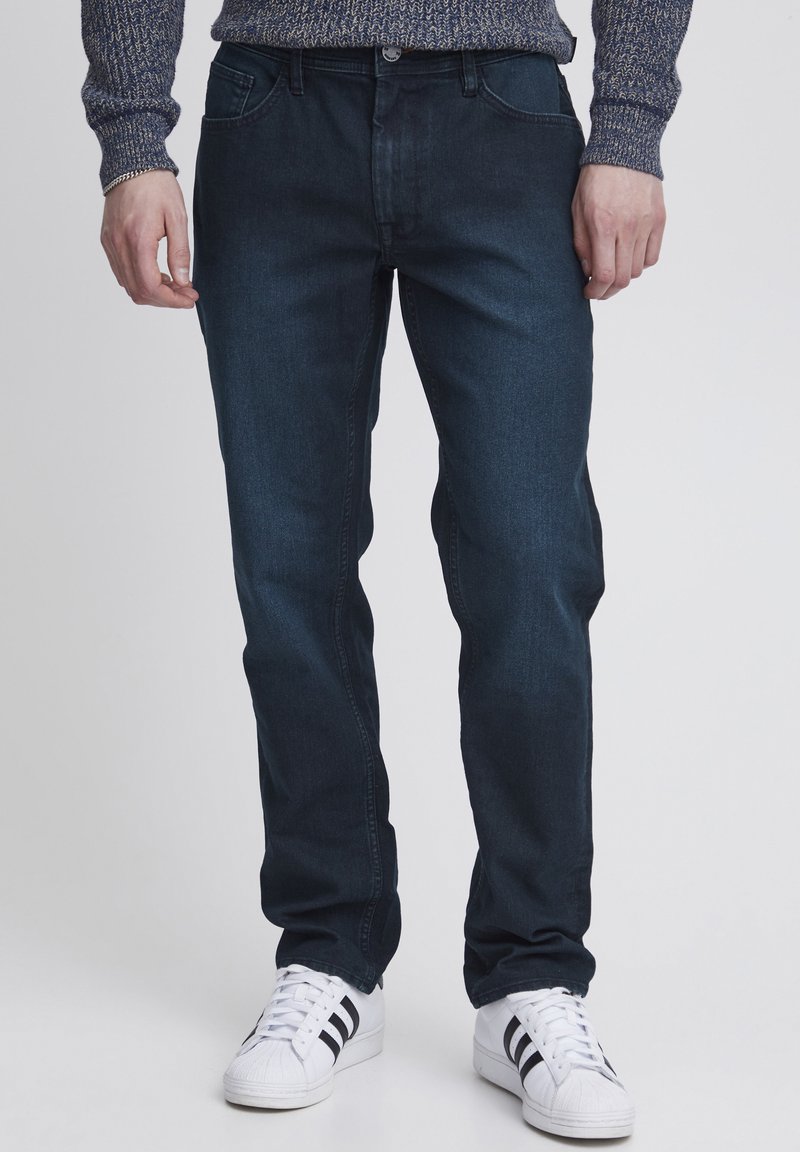 Jeans in denim blu scuro con una vestibilità dritta classica, cinque tasche e zip. Indossati con sneakers bianche con strisce nere.