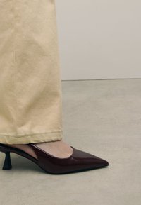Chaussure à talon aiguille en cuir verni bordeaux avec un bout pointu et un talon bloc unique, associée à un pantalon beige légèrement court.