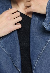 Jeansjacka i ljusblå färg med en texturerad svart topp under. Funktioner inkluderar långa ärmar och sömnadsdetaljer längs sömmarna.