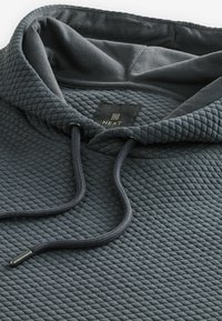 Donkergrijze gewatteerde hoodie met een gestructureerd oppervlak, verstelbaar trekkoord en een zwart label met gouden branding aan de binnenkant van de kraag.