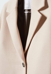 Manteau beige clair au design ajusté, avec des revers pointus et un unique bouton noir. Texture douce et doublure intérieure minimaliste.