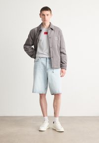 Veste en velours côtelé gris avec l'inscription "HUGO", superposée à un t-shirt gris clair orné d'un logo rouge, associée à un short en denim bleu clair et des baskets blanches.