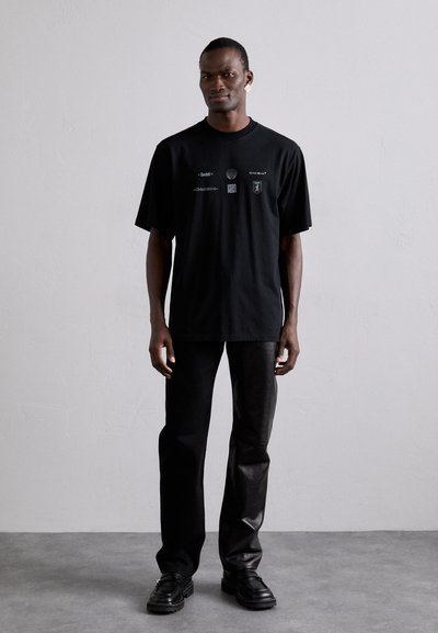 T-shirt noir oversize avec impression graphique sur le devant, associé à un pantalon en cuir noir et des chaussures noires imposantes. Design simple et minimaliste.