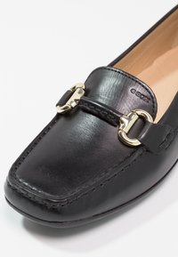 Mocassin en cuir noir avec boucle métallique dorée et coutures apparentes, avec le nom de la marque Geox embossé sur la tige.