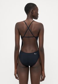 Svart én-delt svømmetøy med racerback-design og kryssede stropper. Har en glatt, jevn tekstur og en liten hvit logo på baksiden.