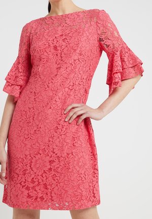 Kobieta w koralowo-różowej koronkowej sukience z warstwowymi falbanami na rękawach do łokcia, ręka oparta na biodrze, na jednolitym jasnym tle.