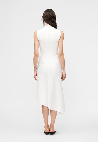 Γυναίκα με μακριά σκούρα μαλλιά, ντυμένη με ασύμμετρο midi φόρεμα χωρίς μανίκια σε λευκό χρώμα και μαύρα παπούτσια με τακούνι kitten, στέκεται μπροστά σε απλό φόντο.