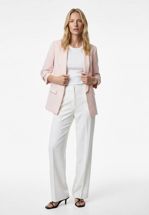 Donna che indossa un blazer rosa chiaro sopra una maglietta bianca con pantaloni bianchi a gamba larga e tacchi aperti neri, in piedi davanti a uno sfondo bianco uniforme.