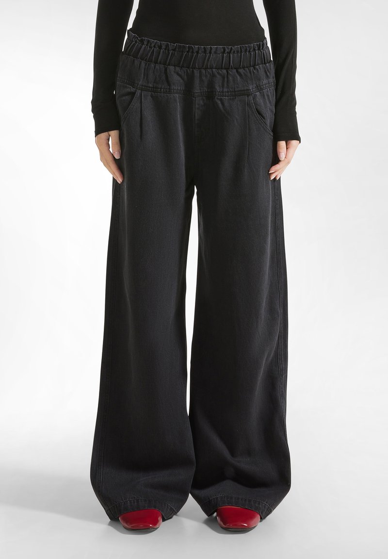 Pantaloni neri a gamba larga in denim, con cintura elastica, due tasche frontali e una texture liscia. I fondi sono leggermente svasati.