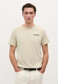 Beige Baumwoll-T-Shirt mit Rundhalsausschnitt und kurzen Ärmeln. Auf der linken Vorderseite befindet sich der dunkelblaue Schriftzug "Scalpers. Films". Kombiniert mit einem schwarzen Gürtel und hellen Hosen.