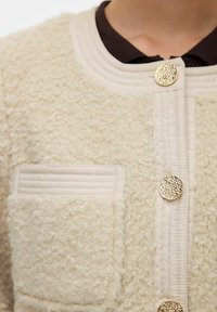 Texturovaná krémová bunda se žebrovaným límcem, dekorativními zlatými knoflíky a boční kapsou. Vyrobena z měkké, chlupaté tkaniny.