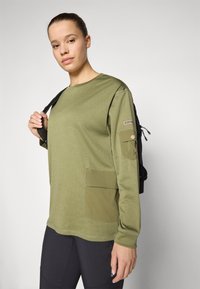 Oliven grønn langermet skjorte med en teksturert finish, sidelomme og et logo på armen. Stilt med svarte leggings og en svart ryggsekk.