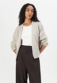 Light beige knit cardigan with open front, ribbed texture, and slightly oversized sleeves, paired with a white fitted top and dark trousers.
