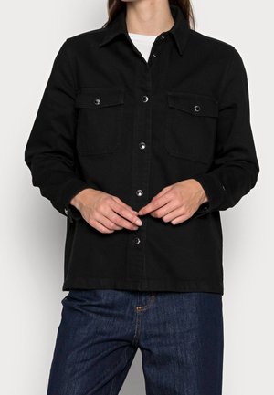 Kobieta w czarnej kurtce zapinanej na guziki z dwiema kieszeniami na piersi, w ciemnoniebieskich dżinsach, stojąca na tle jednolitego białego tła.