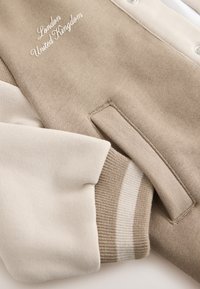 Sweatshirt beige et crème avec broderie "London United Kingdom." Comprend des poignets côtelés et une poche latérale, avec une texture lisse.
