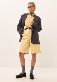 Šedý oversized blejzr, žluté polo, žluté kraťasy, černý opasek, šedé ponožky, černé mokasíny; hladké textury a dobře padnoucí střih.